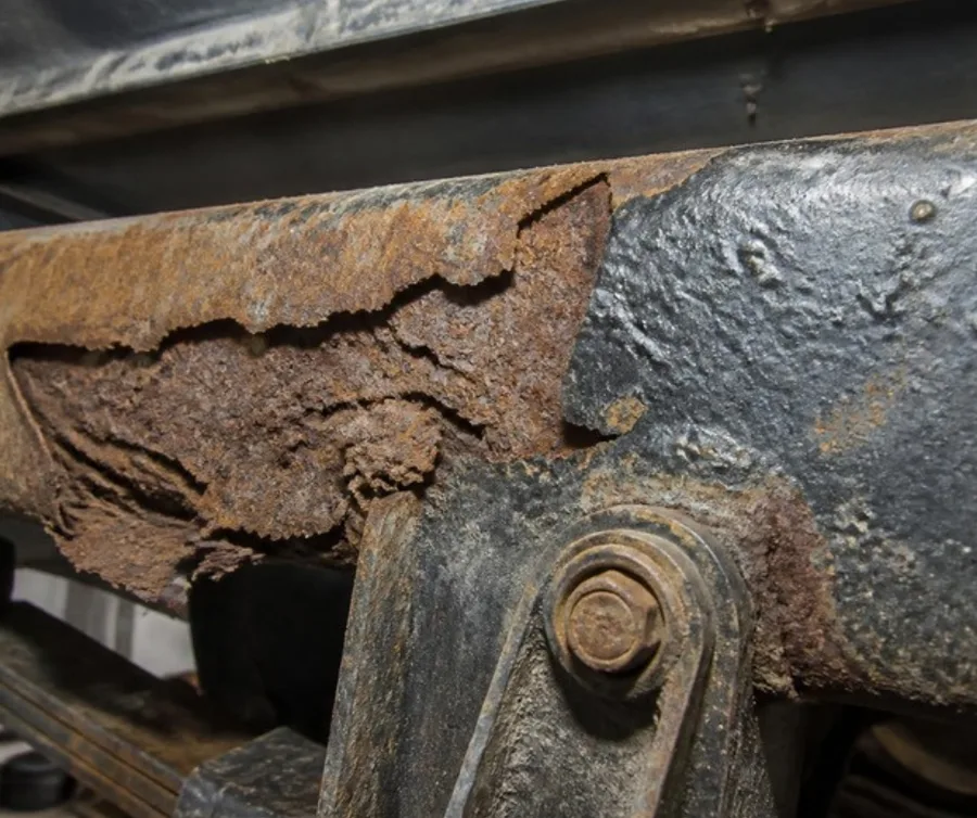 Thick scale rust flaking off a truck frame rail near the rear spring hanger
